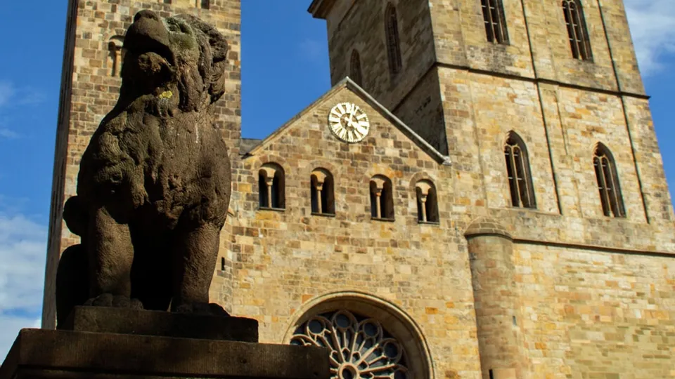 Steinerne Skulptur eines Löwen vor einer historischen Kirche mit zwei Türmen und einer großen Uhr.