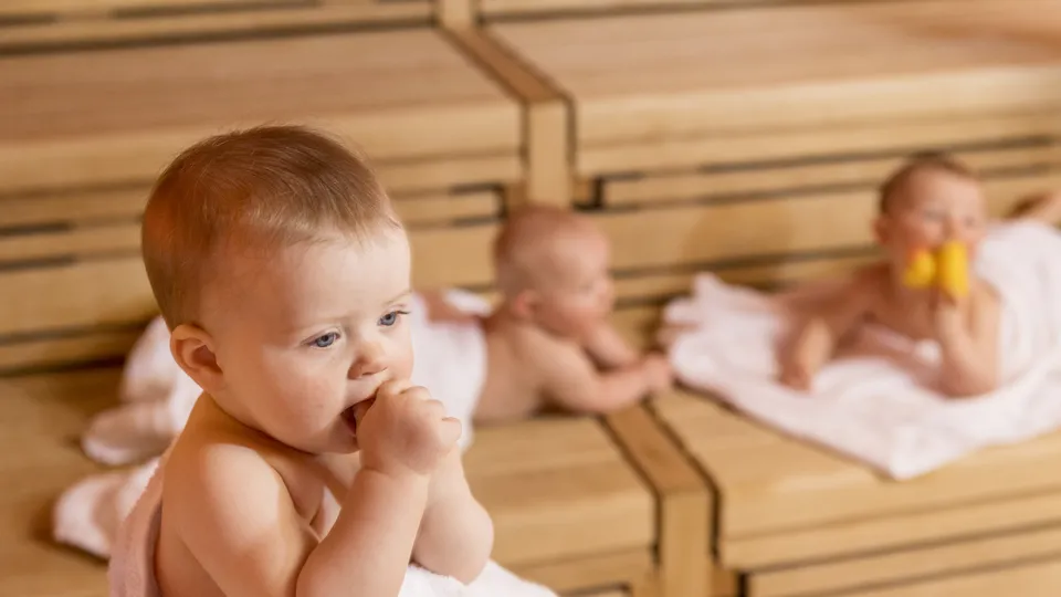 Drei Babys entspannen auf Holzbänken in einer Sauna; eines kaut auf einem Handtuch, zwei ruhen dahinter.