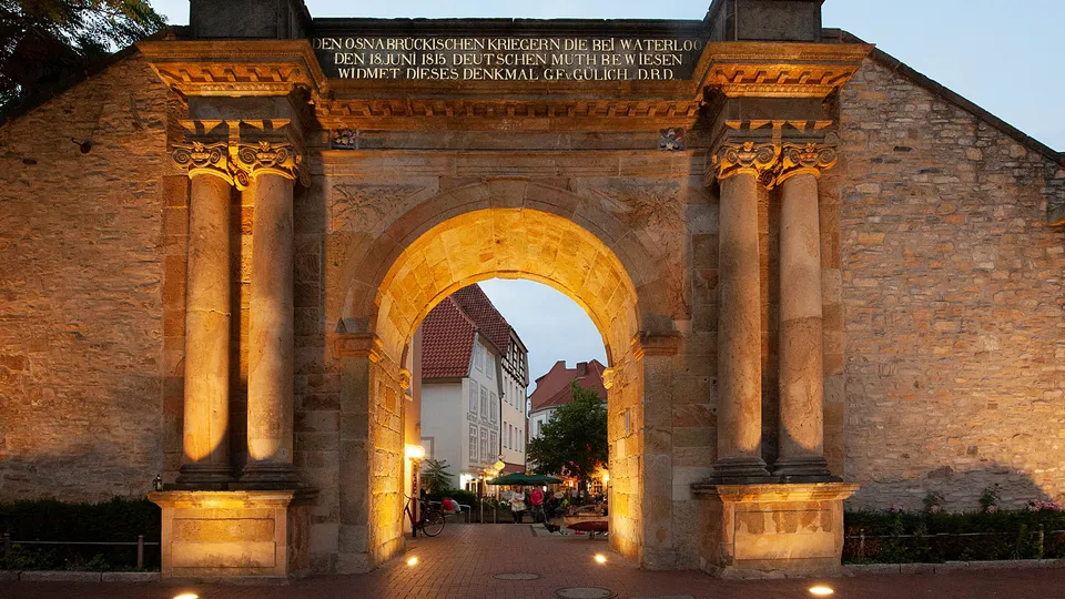 Historisches Tor mit eindrucksvoller Beleuchtung am Abend, Passanten flanieren durch den Eingang.