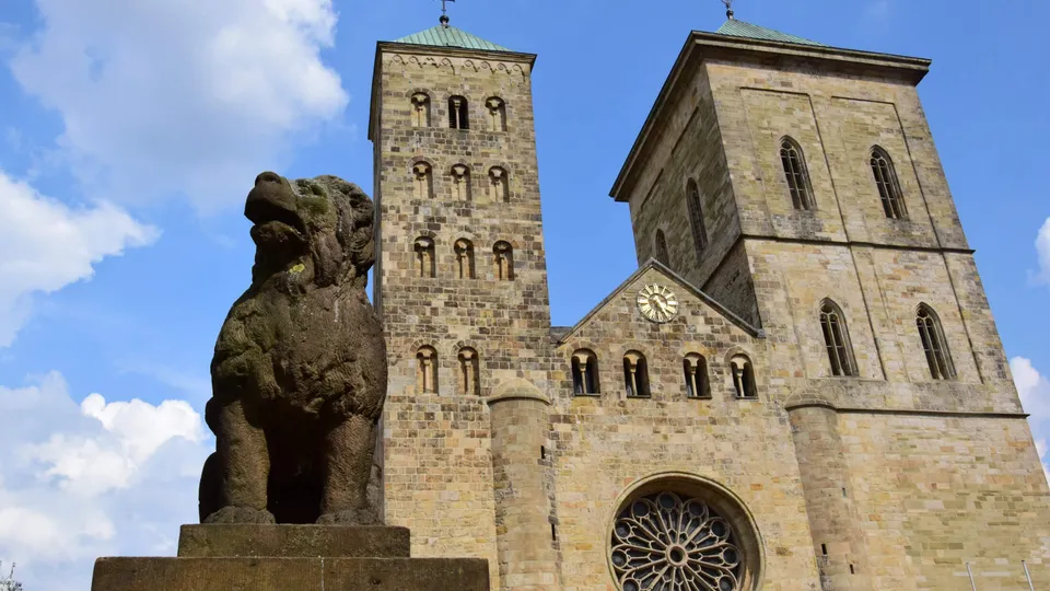 Löwenpudel-Skulptur aus Stein mit zwei Türmen des Osnabrücker Doms und blauem Himmel im Hintergrund.